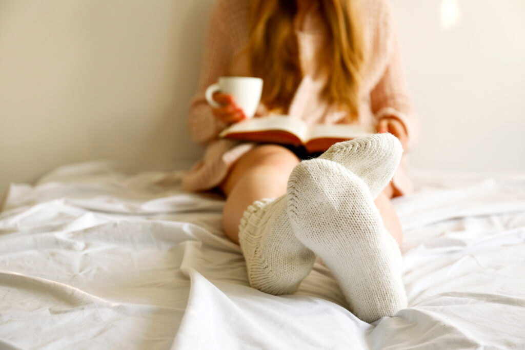 Lazy Afternoon Concept. Close Up Shot Of Woman Sitting In Bed Leaning On The White Wall In Morning Light, By The Window Reading Old Book, Having Cup Of Cappuccino Coffee. Background, Copy Space